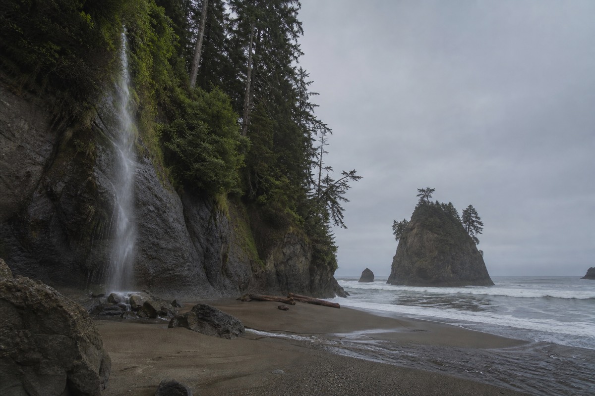 A waterfall spilling over a sandstone bluff onto the sand at Mystic Beach on the Juan de Fuca coast, with forest crowding the cove